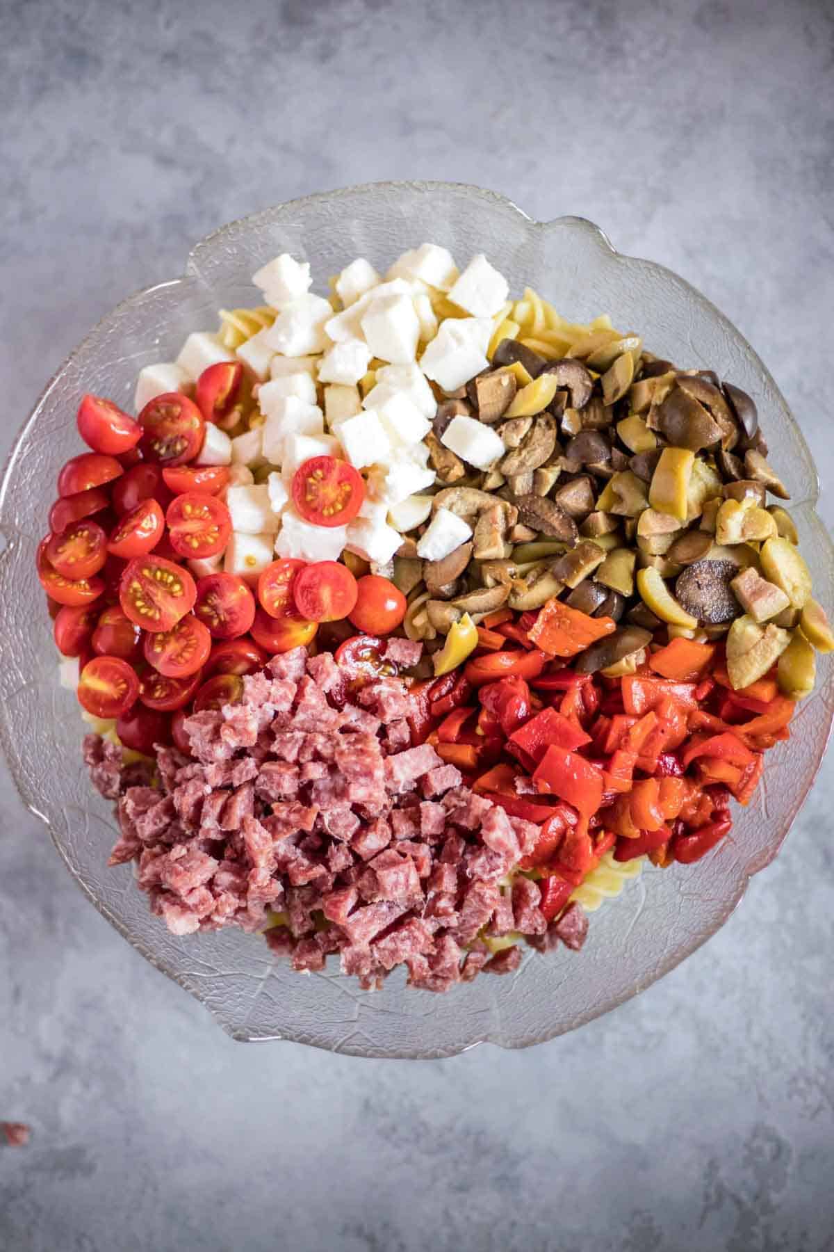 A salad bowl with gluten-free rotini pasta, cherry tomatoes, mozzarella balls, olives, roasted red pepper and salami.