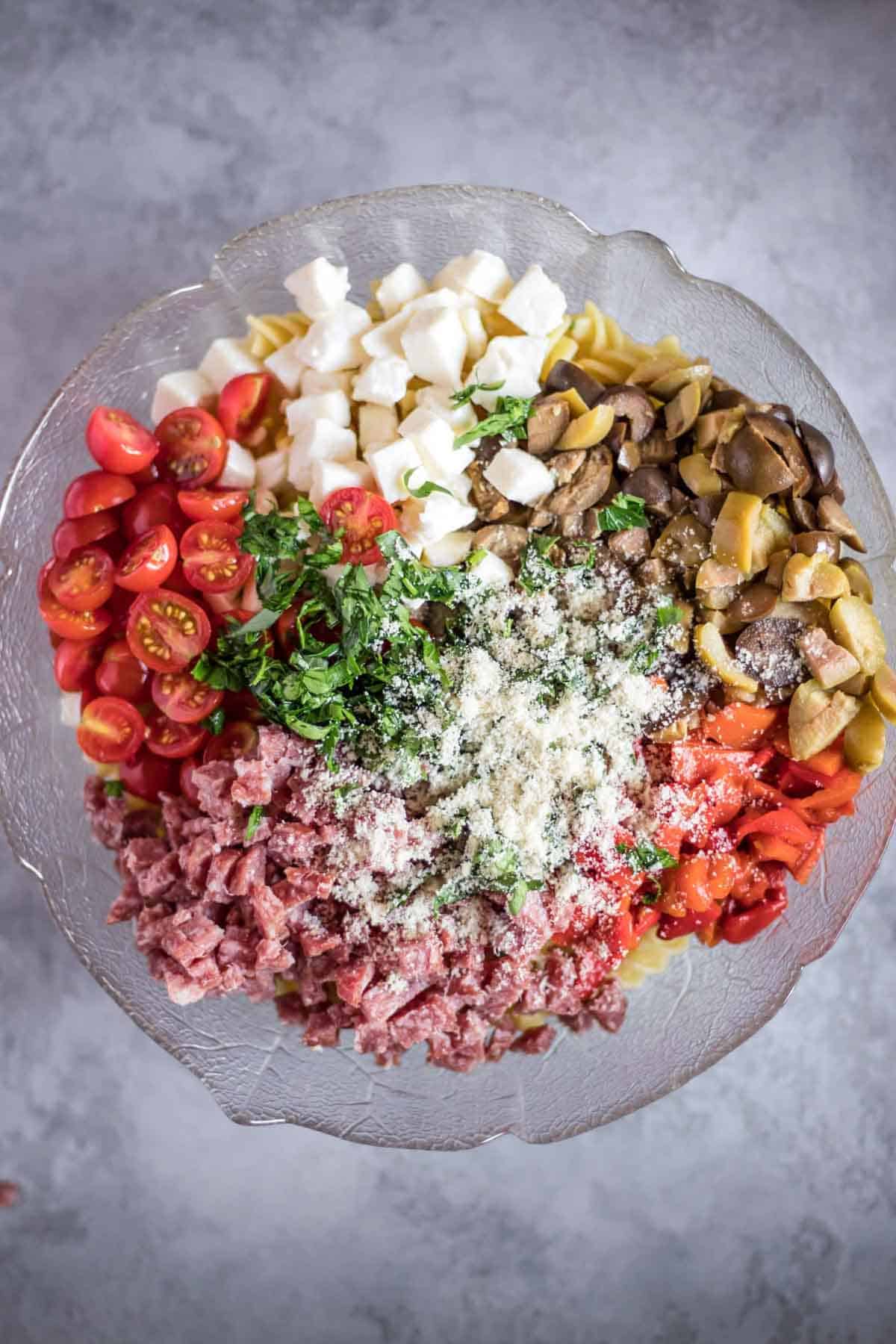A salad bowl with gluten-free rotini pasta, cherry tomatoes, mozzarella balls, olives, roasted red pepper, salami, parsley and parmesan cheese.