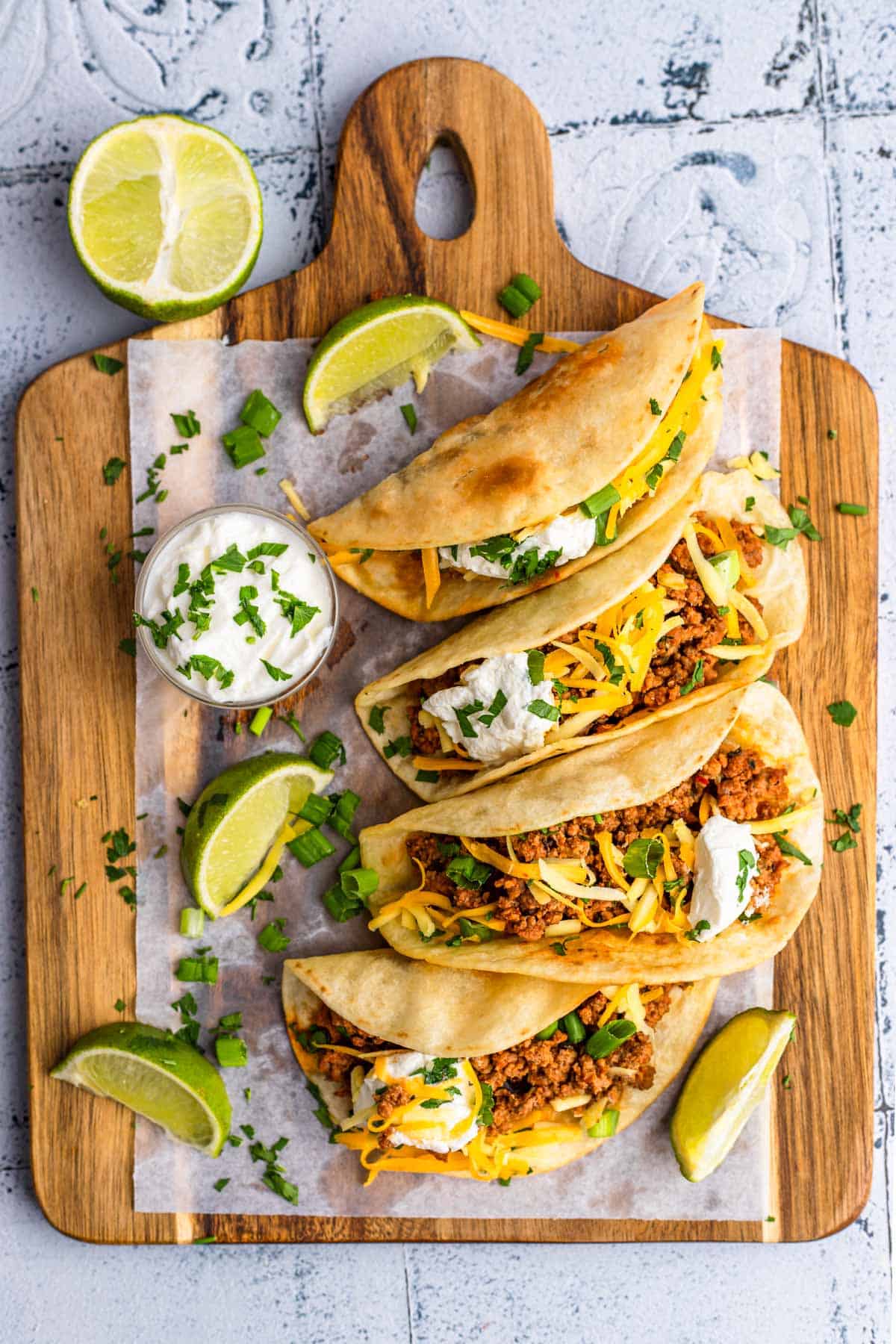 Four tacos plated on a wooden board, a small bowl of sour cream and lime wedges