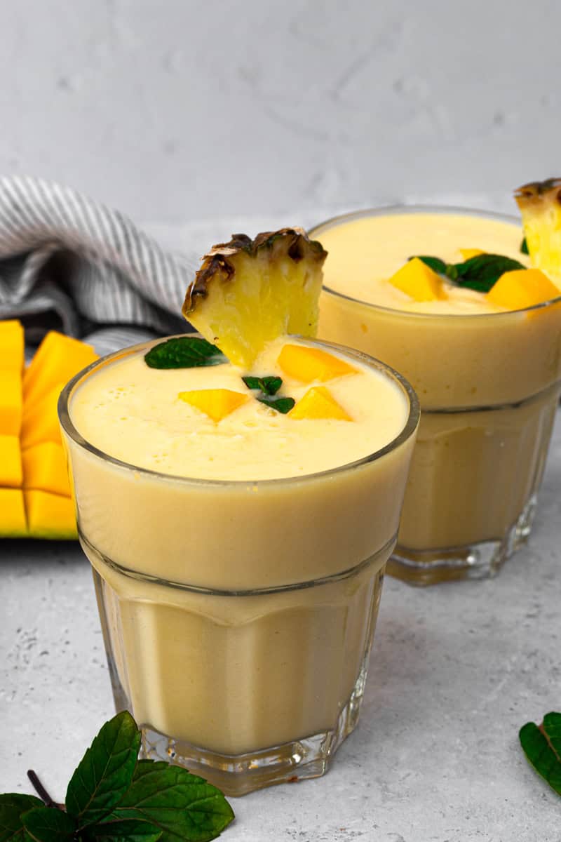 tall glass full of mango smoothie mango chunks and a slice of pineapple on the rim of the glass