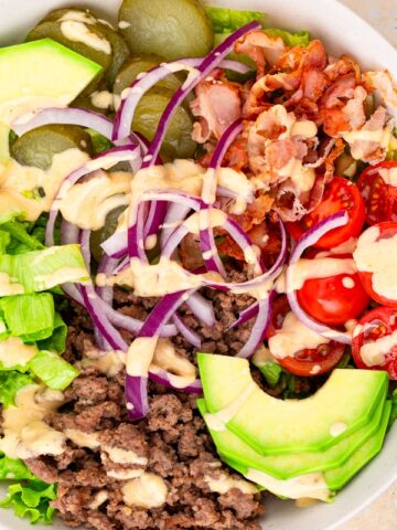overhead view of burger bowl topped with burger sauce and avocado, sliced red onions, cherry tomatoes, pickles, cooked bacon and lettuce.
