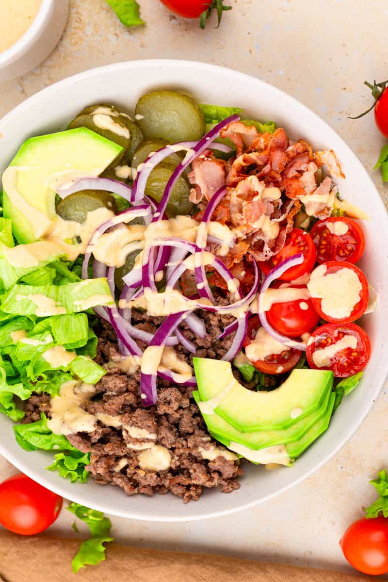 overhead view of burger bowl topped with burger sauce and avocado, sliced red onions, cherry tomatoes, pickles, cooked bacon and lettuce.